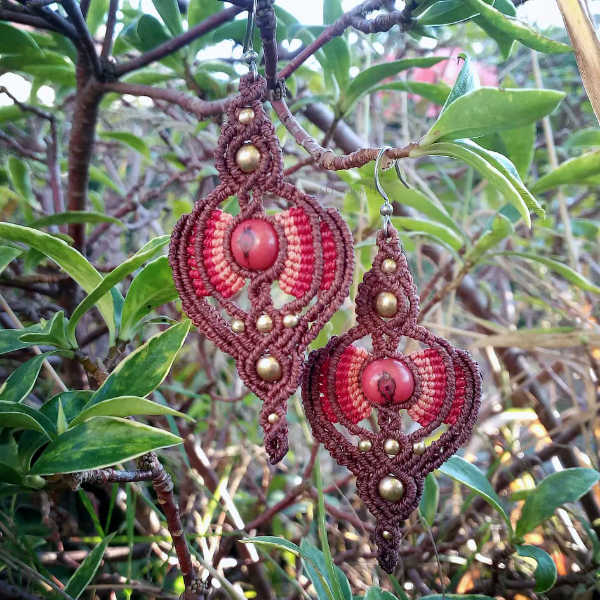 Boucles d'oreilles en micro-macramé, effet rayonnant autour d'une graine d'açaï rouge, fils marron acajou, rouge et terracotta, perles en laiton, arrière plan végétation