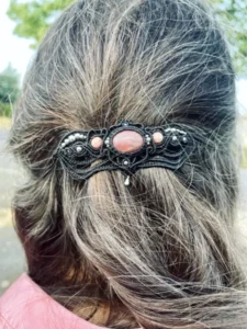 Barrette à cheveux en micro-macramé noir, cabochon et perles rhodonite, finitions acier inoxydable, motifs baroques, vue portée cheveux châtain gris, col vieux rose, arrière plan végétation