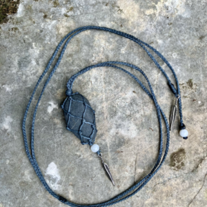 Pendentif en micro-macramé gris anthracite, galet naturel serti en filet, perles en pierres de Lune et plumes en métal, posé sur une pierre plate