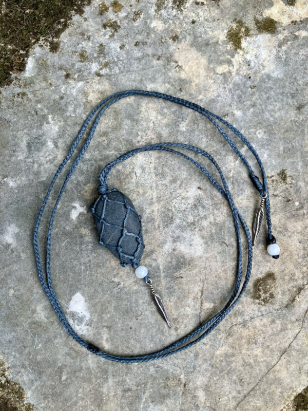 Pendentif en micro-macramé gris anthracite, galet naturel serti en filet, perles en pierres de Lune et plumes en métal, posé sur une pierre plate