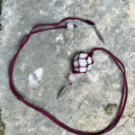 Pendentif en micro-macramé violet prune, galet naturel serti en filet, perles en quartz rose et plumes en métal, posé sur une pierre plate