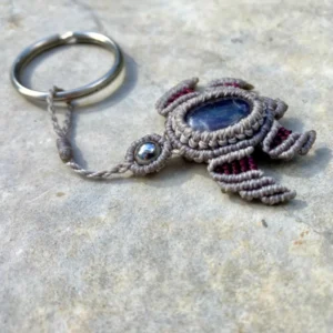 Porte-clefs en micro-macramé 3D, vue de 3/4, tortue marine gris et prune, carapace : cabochon améthyste, tête : perle en acier inoxydable, anneau en acier inoxydable