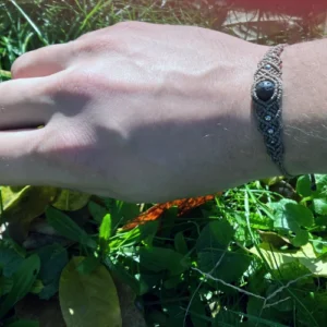 Bracelet en micro-macramé marron cèdre, perles en pierre de lave et acier inoxydable, motif entrelacs, vue porté, zoom sur la main arrière plan végétation