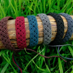 6 bracelets en micro-macramé acajou, bourgogne, bleu sarcelle, cèdre, noir et gris anthracite, motif double entrelacs, sur un support bois, arrière plan végétation