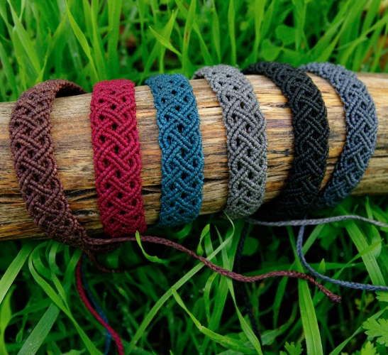 6 bracelets en micro-macramé acajou, bourgogne, bleu sarcelle, cèdre, noir et gris anthracite, motif double entrelacs, sur un support bois, arrière plan végétation