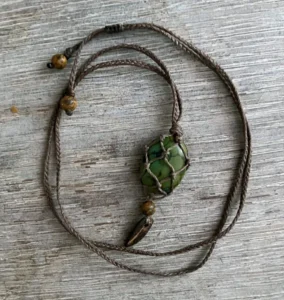 Collier en micro-macramé marron cèdre, pierre roulée inconnue sertie en filet, perles jaspe paysage et morceau de noix de coco, vue posé sur une table en bois