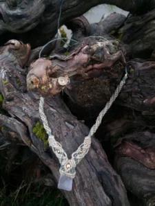 Collier en micro-macramé fils de lin ciré beige et khaki, cristal de roche et perles en laiton, exposé sur des ceps de vignes entassés