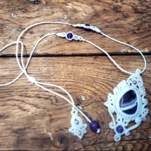 Pendentif en micro-macramé blanc, cabochon et perles en améthystes, perles en acier inoxydables, motifs feuilles, entrelacs, baroque, sur table en bois