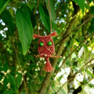 Petite suspension hiboux en micro-macramé orange cannelle, yeux en perles de bois teintées vert, petite branche, arrière plan végétation