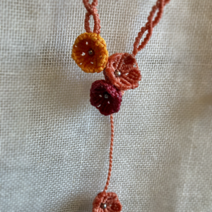 Zoom sur les fleurs d'un collier sautoir asymétrique Trio floral suspendu devant un tissu beige. Fils orange terracotta, topaze et rose framboise, perles en acier inoxydable