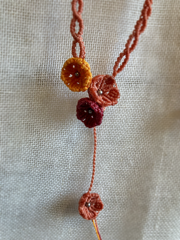 Zoom sur les fleurs d'un collier sautoir asymétrique Trio floral suspendu devant un tissu beige. Fils orange terracotta, topaze et rose framboise, perles en acier inoxydable
