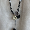 Zoom sur les fleurs d'un collier sautoir asymétrique Trio floral suspendu devant un tissu beige. Fils noir, gris et blanc naturel, perles en acier inoxydable et pierres de Lune