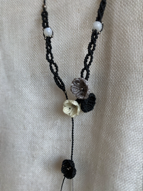 Zoom sur les fleurs d'un collier sautoir asymétrique Trio floral suspendu devant un tissu beige. Fils noir, gris et blanc naturel, perles en acier inoxydable et pierres de Lune
