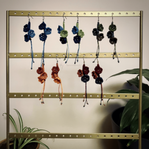 Présentation de 5 paires de boucles d'oreilles Trio floral suspendues sur un support. Arrière-plan beige, plantes.