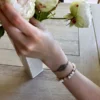 Photo bracelet à motif feuille en micro-macramé beige, porté avec un bracelet de perles blanc et doré, arrière plan vase avec roses et table en bois clair