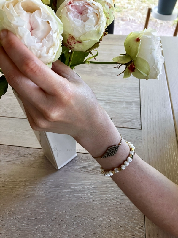 Photo bracelet à motif feuille en micro-macramé beige, porté avec un bracelet de perles blanc et doré, arrière plan vase avec roses et table en bois clair