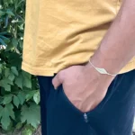 Photo zoom sur bracelet à motif feuille en micro-macramé blanc naturel, porté par un homme debout, main dans la poche, arrière plan feuillage et muret