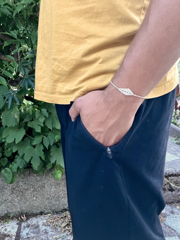 Photo zoom sur bracelet à motif feuille en micro-macramé blanc naturel, porté par un homme debout, main dans la poche, arrière plan feuillage et muret