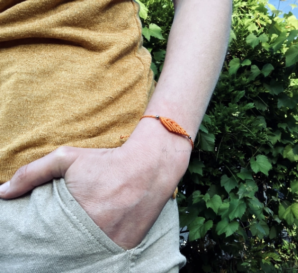 Photo détail d'un bracelet au motif feuille en micro-macramé jaune topaze et perles en acier inoxydable, porté, main dans la poche, végétation en arrière-plan