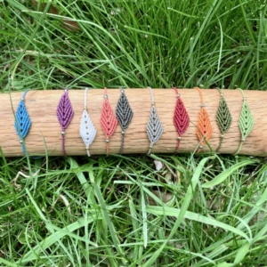 Photo 10 bracelets en micro-macram&eacute; de diff&eacute;rentes couleurs, motif feuille simple, expos&eacute;s sur un support en bois clair, herbe en arri&egrave;re-plan