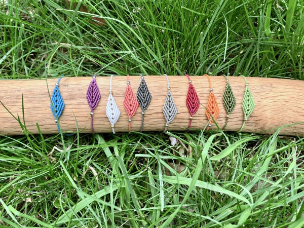 Photo 10 bracelets en micro-macramé de différentes couleurs, motif feuille simple, exposés sur un support en bois clair, herbe en arrière-plan