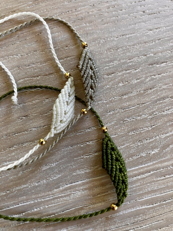 Photo de trois bracelets à motif feuille en micro-macramé blanc naturel, beige et vert olive, posés sur une table en bois clair